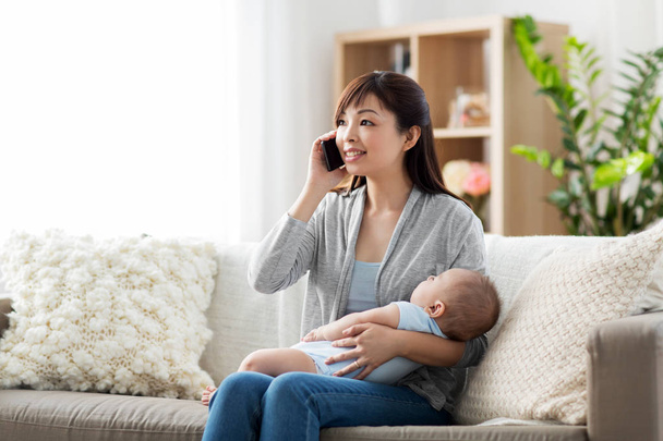 family, technology and motherhood concept - happy smiling young asian mother with sleeping baby calling on smartphone at home - Фото, изображение