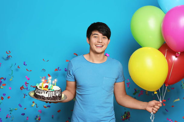 Young man with birthday cake and bright balloons on color background - Фото, изображение