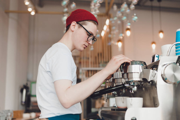 A young man works as a Barista he stands behind the coffee machine and makes coffee - Fotoğraf, Görsel
