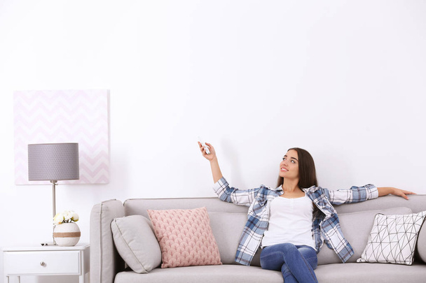 Young woman switching on air conditioner while sitting on sofa near white wall - Φωτογραφία, εικόνα