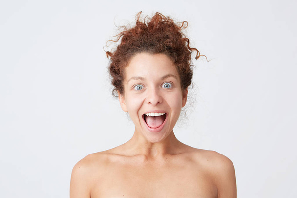 Cropped shot of happy excited curly young woman with opened mouth looking amazed and has healthy skin after applying mask or body cream, takes care of her appearance, stands half naked isolated over white background - Photo, Image