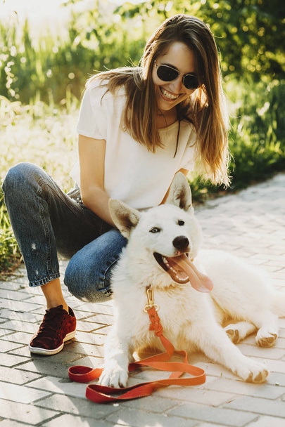 lachende schöne junge Frau in Sommerkleidung und Sonnenbrille, die auf Bunkern sitzt, während ihr weißer Hund auf dem Bürgersteig des Parks liegt, mit herausgestreckter Zunge auf verschwommenem hinterleuchteten Hintergrund - Foto, Bild