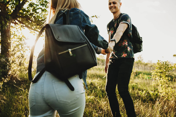 Corte a visão traseira da fêmea em jeans roupas de verão segurando a mão de macho bonito feliz e levando-o com ela de pé na grama verde iluminada pelo sol com árvore
 - Foto, Imagem