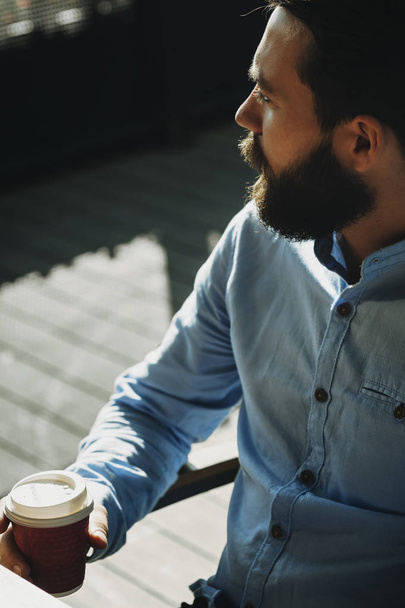 Crop view of bearded male in light shirt sitting outdoors with closed paper cup with tea or coffee and looking away with sunlit pavement on blurred background - Photo, Image