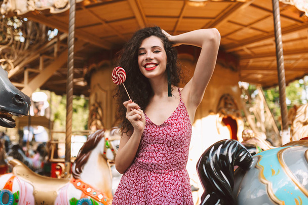 Krásná dáma s tmavými kudrnatými vlasy šaty stojí s lolly pop candy v ruce při šťastně pohledu kamery s rotujícím na pozadí - Fotografie, Obrázek