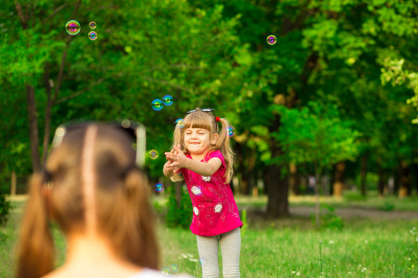Young mother and little daughter playing in park with soap bubbles. Love family, parenthood, childhood - Fotografie, Obrázek