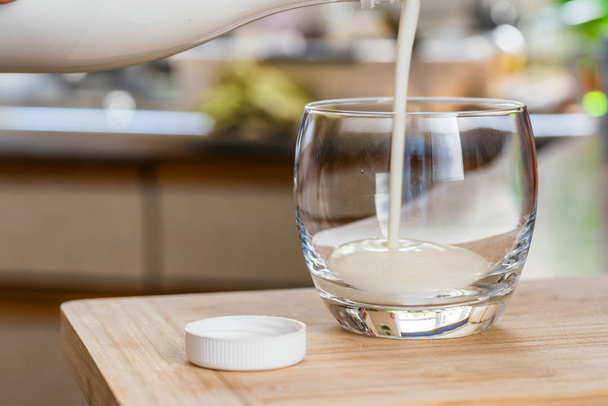 Hand pouring fresh kefir probiotik drink from white bottle into clear glass on kitchen table - Photo, Image
