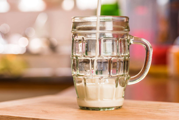 Vista de cerca verter kéfir fresco probiotik bebida en un cuarto de taza de vidrio transparente completo en la mesa de la cocina
 - Foto, Imagen