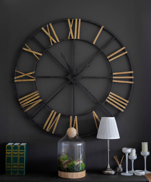 The Big Vintage Steel Clock Roman Numerals on The Black Wall with The Green Vintage Mock up Books, The Plants in The Modern Glass Bottle, White Lamp, White Candles with Candlestick and Aroma Incense Stick in Little Glass Bottle. - Photo, Image