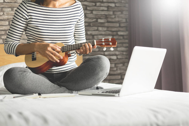 Happy young asian woman playing ukulele sitting on bed in bedroom - Photo, Image