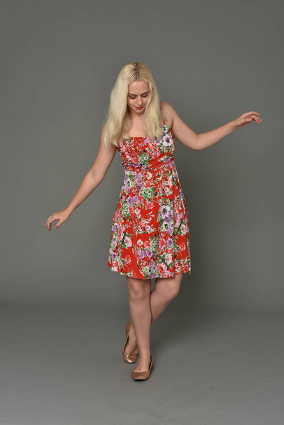 полноразмерный портрет блондинки в красном цветочном платье, стоящая поза на сером фоне студии
. - Фото, изображение
