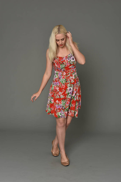 full length portrait of blonde girl wearing red floral dress, standing pose on grey studio background. - Foto, afbeelding