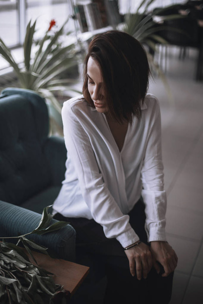 A beautiful girl is sitting on a blue velvet chair and is resting. A woman in black trousers and a white shirt. brunette with short hair. businesswoman in the office. - Фото, зображення