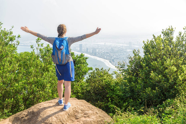 Young female tourist raising hands and enjoying beautiful view and fresh air on top of mountain among green bushes. Girl with blue backpack. A coastal city is visible in background. - Foto, imagen