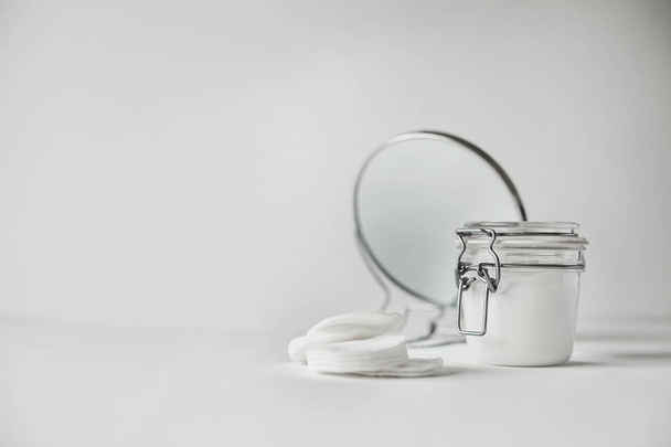 Красивая минималистская концептуальная вся белая композиция - хлопчатобумажные прокладки, прозрачная банка и круглое зеркало, все белое, с металлическими деталями
 - Фото, изображение
