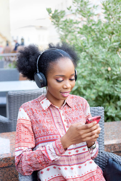 Chica afro negro en vestido étnico con auriculares en el cuello sentado en la cafetería al aire libre, escribiendo en el teléfono inteligente y sonriendo
. - Foto, imagen