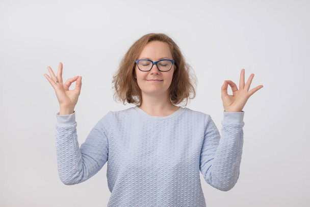jeune femme souriante européenne en pull bleu et des lunettes modernes méditant, tenant ses mains dans le geste de yoga, se sentant calme et positive avec les yeux fermés
 - Photo, image