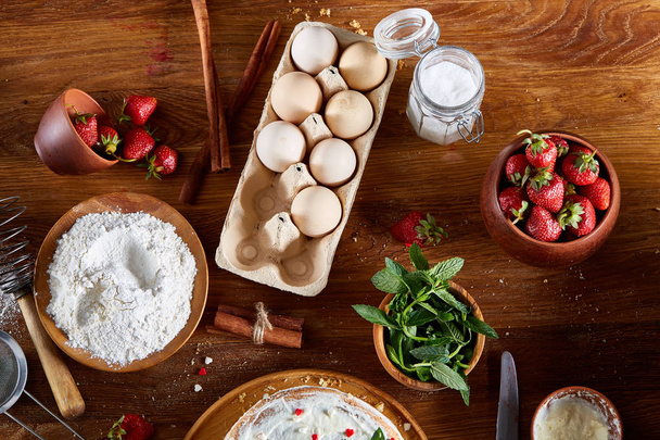 Strawberry tart covered with whipped sour cream surrounded by organic ingredients over wooden background, flat lay. - Foto, imagen