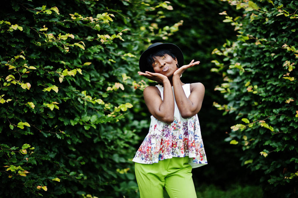 Geweldige Afro-Amerikaanse model vrouw in groene broek en zwarte hoed gesteld in het park. - Foto, afbeelding