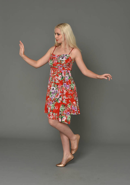full length portrait of blonde girl wearing red floral dress, standing pose on grey studio background. - Photo, image