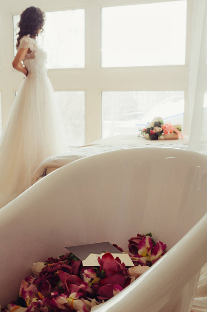 Un baño blanco con flores rojas y una carta en el sobre, con novia borrosa abotonando su vestido en el fondo mirando a la ventana, su ramo de bodas en una cama blanca junto a ella
 - Foto, Imagen