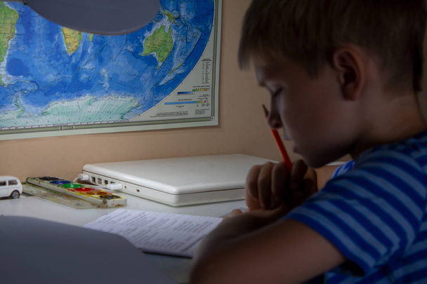 家庭教育。放課後の家の仕事。伝統的な白いメモ帳の紙に手で英語のテストを書くペンを持つ少年.  - 写真・画像