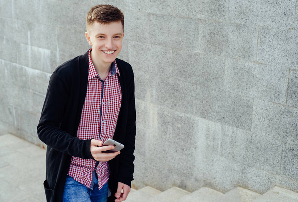 Elegante joven sonriente con camisa a cuadros y jeans subiendo escaleras y utilizando el teléfono inteligente al aire libre. Estudiante con smartphone subiendo escaleras, espacio libre
 - Foto, imagen