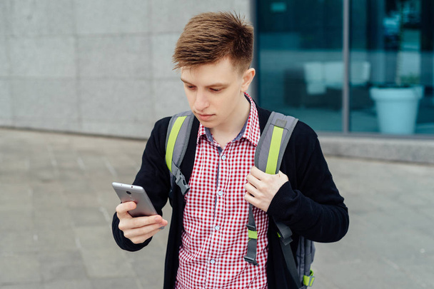 Stylish young man in plaid shirt with backpack walking in the city and using cell phone outdoors. Student with smartphone walking  - Foto, Imagen