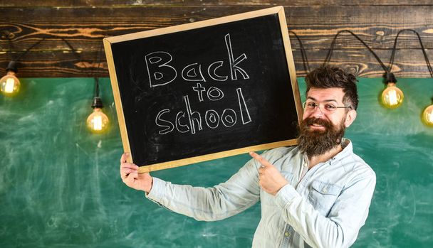 Teacher in eyeglasses holds blackboard with title back to school. Hiring teachers concept. Man with beard and mustache on happy face welcomes colleagues, chalkboard on background - Photo, Image