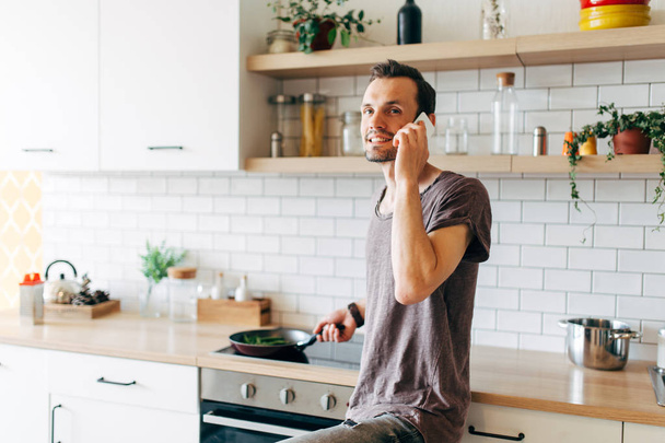 Portrait of brunet man with frying pan in his hands talking on phone in kitchen - 写真・画像