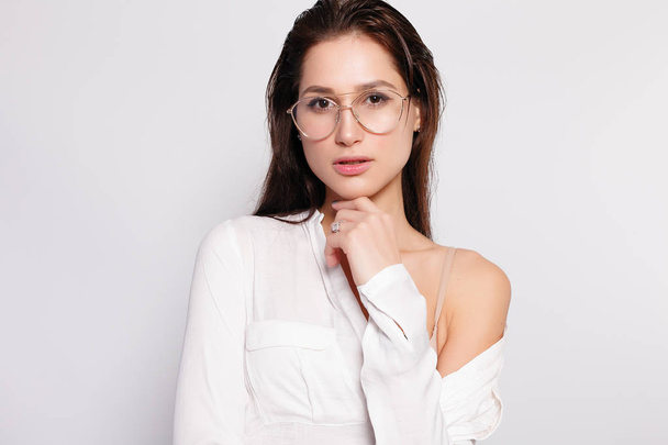 Pretty young girl in white blank t-shirt, wearing glasses, empty wall, studio portrait. Smiling brunette woman in eyeglasses - Фото, изображение