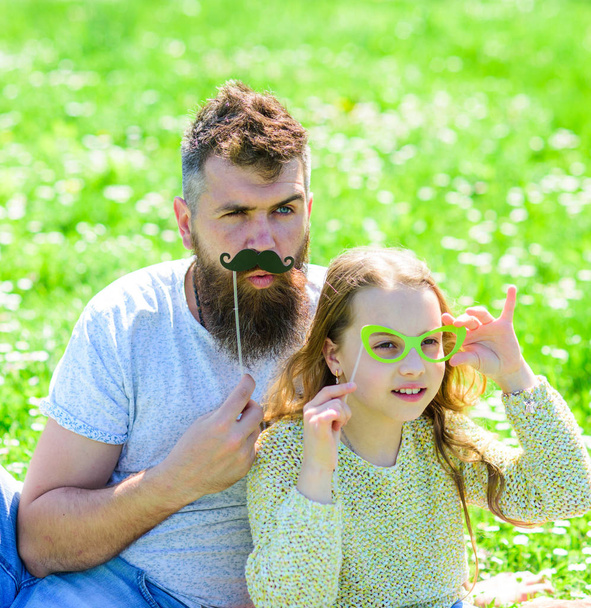 Father posing with mustache and child posing with eyeglasses photo booth attribute. Dad and daughter sits on grassplot, grass on background. Detectives concept. Family spend leisure outdoors - Foto, afbeelding
