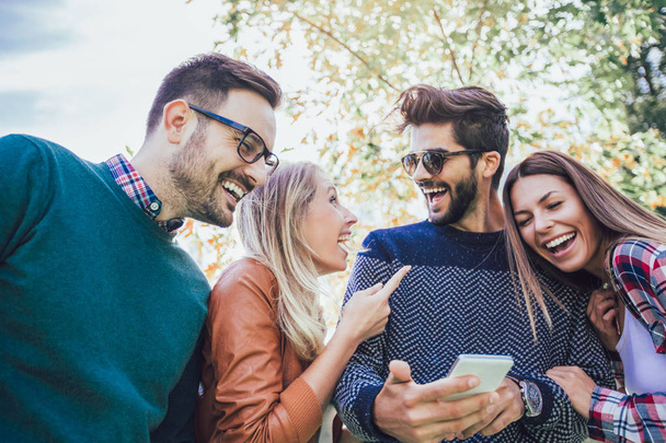 Image de quatre jeunes amis souriants heureux marchant à l'extérieur dans le parc tenant un téléphone intelligent
 - Photo, image