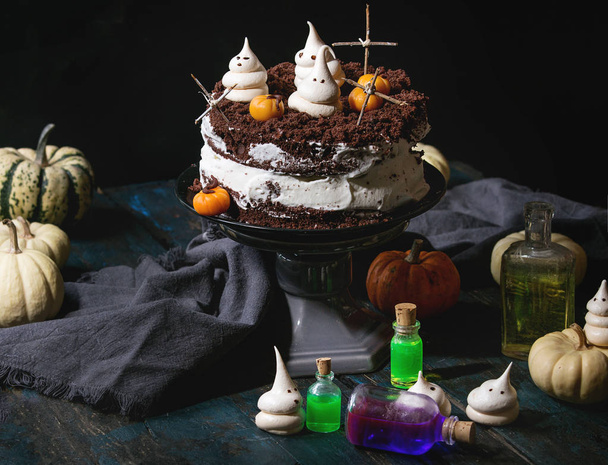 Хэллоуинский сладкий стол с шоколадным тортом на кладбище, марципаном и декоративными тыквами, безе призраки, ядовитые бутылки на черном деревянном столе
. - Фото, изображение