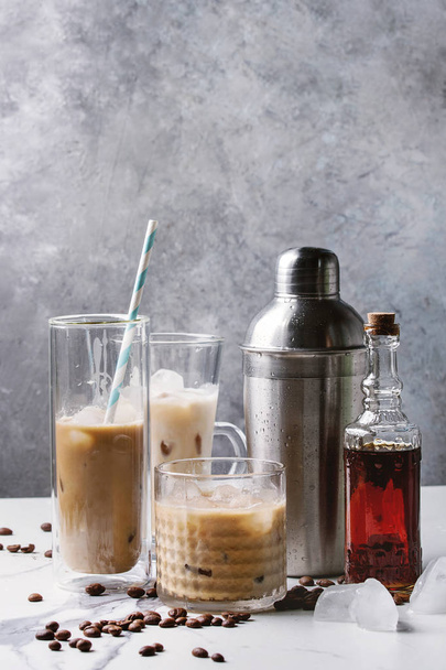 Iced coffee cocktail or frappe with ice cubes and cream in different glasses with silver shaker, bottle of rum, coffee beans around on white marble table with grey concrete wall at background. - Foto, imagen