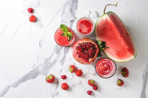 Three different red fruit berry watermelon, strawberry, raspberry, pomegranate cocktails or smoothies in glasses with crushed ice, fresh mint, ingredients above over white marble background. Flat lay - Foto, afbeelding