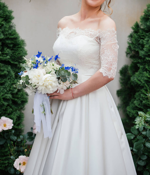 Beautiful bride in wedding dress with wedding bouquet of peonies, blue flowers and greenery outdoors - Photo, Image