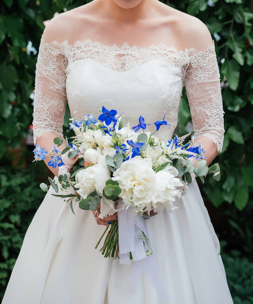 Beautiful bride in wedding dress with wedding bouquet of peonies, blue flowers and greenery outdoors with green leaves in blur on background - Foto, afbeelding