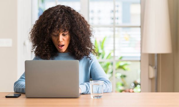 Αφρικανική αμερικανική γυναίκα που χρησιμοποιούν φορητό υπολογιστή στο σπίτι φοβισμένος σε κατάσταση σοκ με πρόσωπο έκπληξη, φοβάται και ενθουσιασμένος με φόβο έκφραση - Φωτογραφία, εικόνα