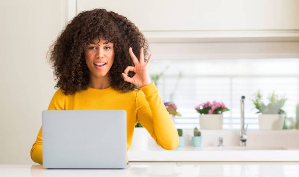 African american woman using computer laptop at kitchen doing ok sign with fingers, excellent symbol - 写真・画像