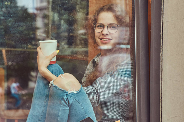Souriant jeune rousse fille bouclée portant des vêtements décontractés et des lunettes assis sur un rebord de fenêtre avec un café à emporter
. - Photo, image