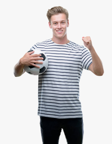 Young handsome blond man holding soccer ball screaming proud and celebrating victory and success very excited, cheering emotion - Foto, immagini