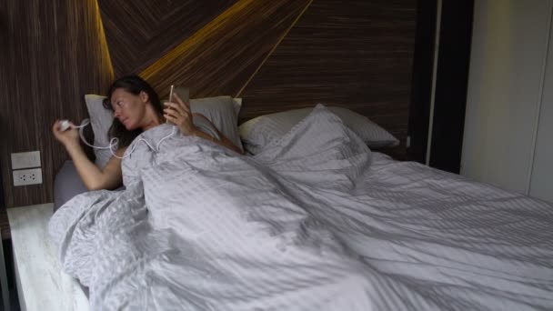 A woman lies in bed and includes phone in charger - Imágenes, Vídeo