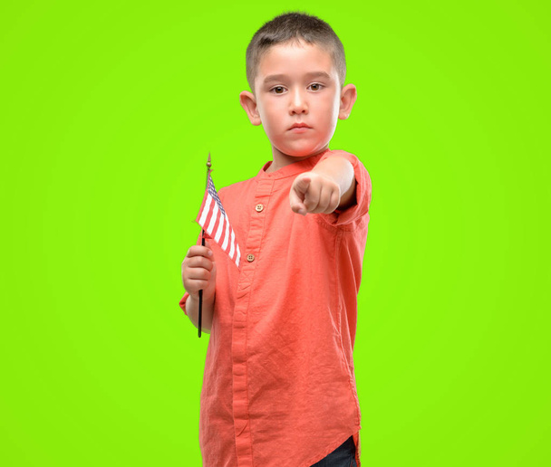 Dark haired kindje houden van de vlag van de Verenigde Staten met de vinger te wijzen naar de camera en u, hand teken, positieve en zelfverzekerd gebaar van de voorkant - Foto, afbeelding