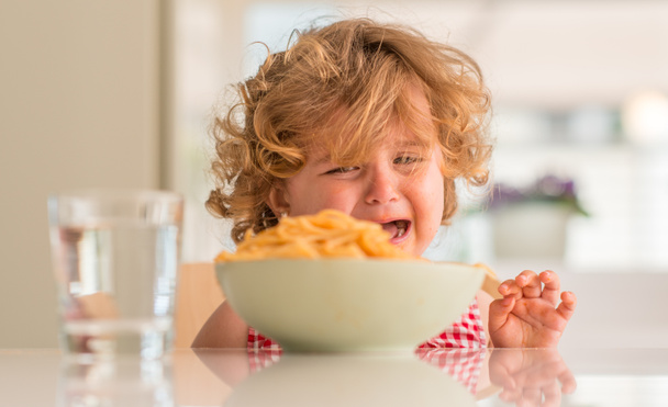 Όμορφο ξανθό παιδί τρώει μακαρόνια με τα χέρια να κλαίνε με ξέσπασμα στο σπίτι. - Φωτογραφία, εικόνα