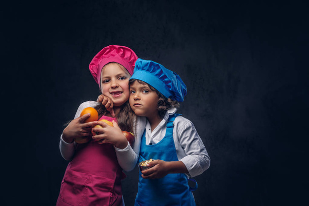 Cute cook couple. Little boy with brown curly hair dressed in a blue cook uniform and beautiful schoolgirl dressed in a pink cook uniform, poses with cookware and fruits at a studio. Isolated on a dark textured background. - Фото, изображение