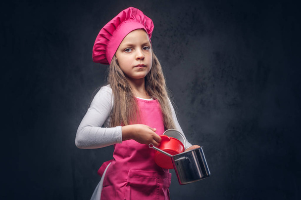 Cute beautiful schoolgirl dressed in a pink cook uniform holds cookware at a studio. Isolated on the dark textured background. - Φωτογραφία, εικόνα
