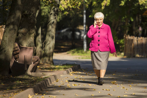 A beautiful senior woman is walking and talking on the phone in a city park on sunny day. - Photo, image