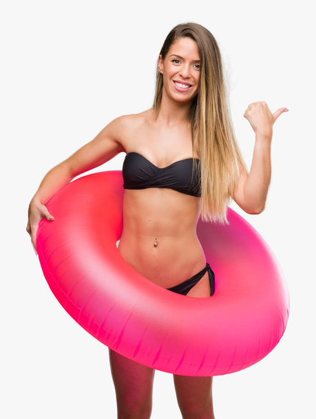 Gyönyörű fiatal nő a bikini gazdaság rózsaszín úszó, mosolygós boldog arc, kéz és az ujjak felfelé mutató - Fotó, kép