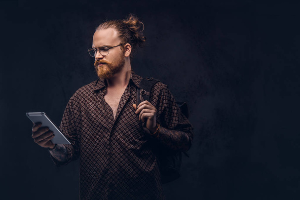Portrait of a redhead hipster student in glasses dressed in a brown shirt, holds a backpack and digital tablet, posing at a studio. Isolated on the dark background. - Photo, Image